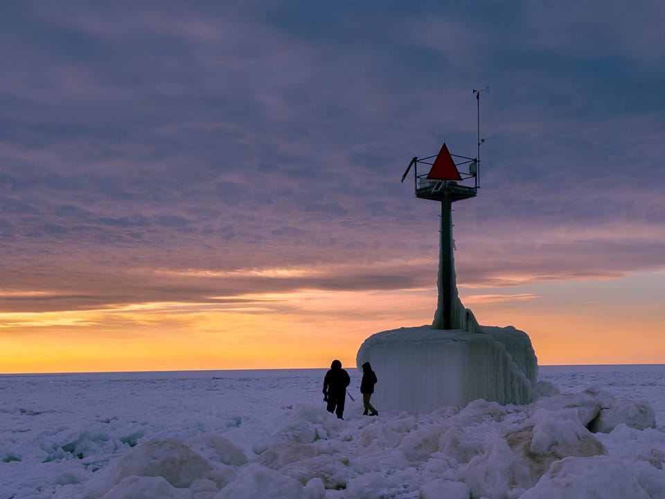 Two silhouetted people stand in front of a frozen pier light with an expansive sky and sunset in the background.